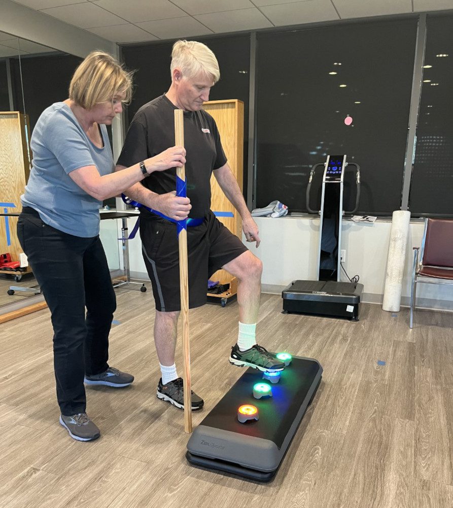 A physical therapist guides a patient in how to step up and touch a light with his foot to improve his reaction timing. 